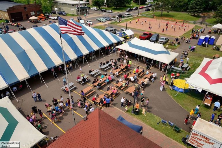 Crowd at a festival in Powderhorn Village, Michigan, with tents, picnic tables, and a volleyball game.
