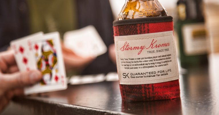 Stormy Kromer beer koozie on a bar with playing cards. "Stormy Kromer, True. Since 1903. Guaranteed for life.