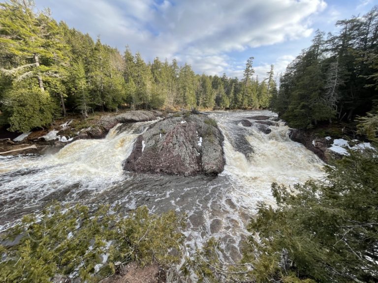 Scenic waterfalls near Powderhorn Village cascade around a large rock, surrounded by evergreen trees.