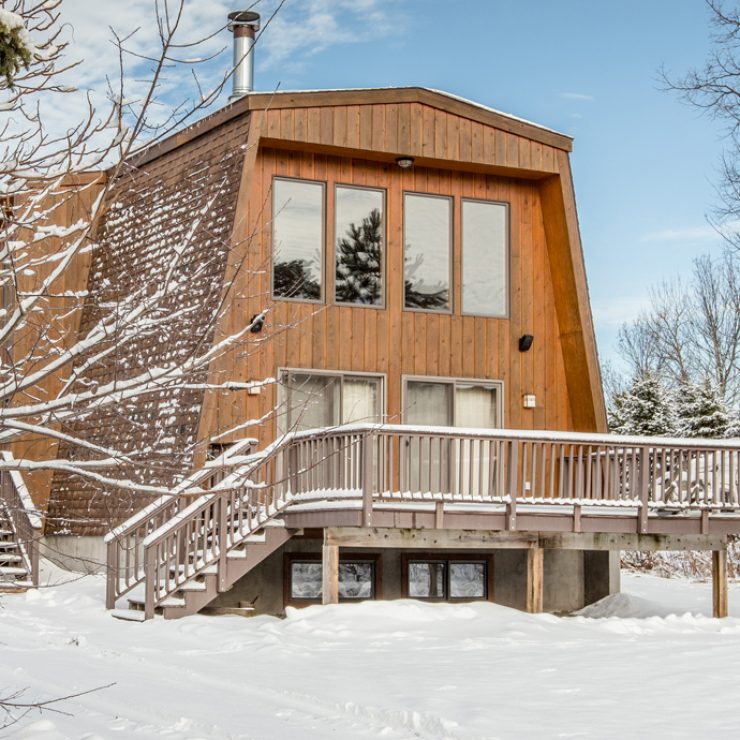 Winter cabin with snow-covered deck and large windows reflecting trees.