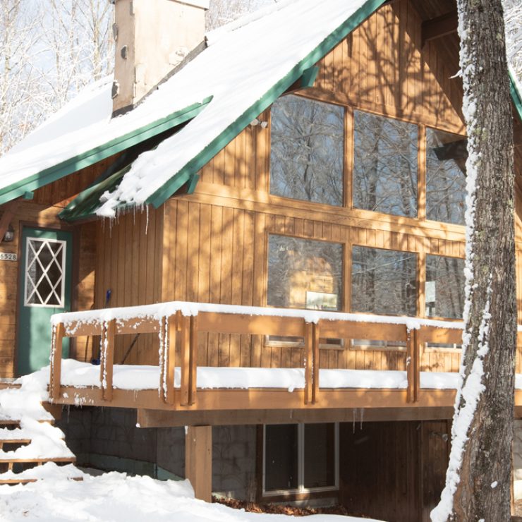 Snow-covered wooden cabin with green trim and a deck, nestled in a winter forest.