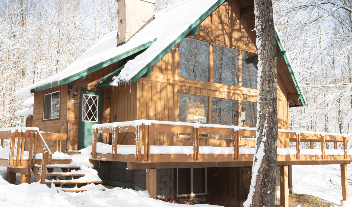 Snow-covered wooden cabin with green trim and a deck, nestled in a winter forest.
