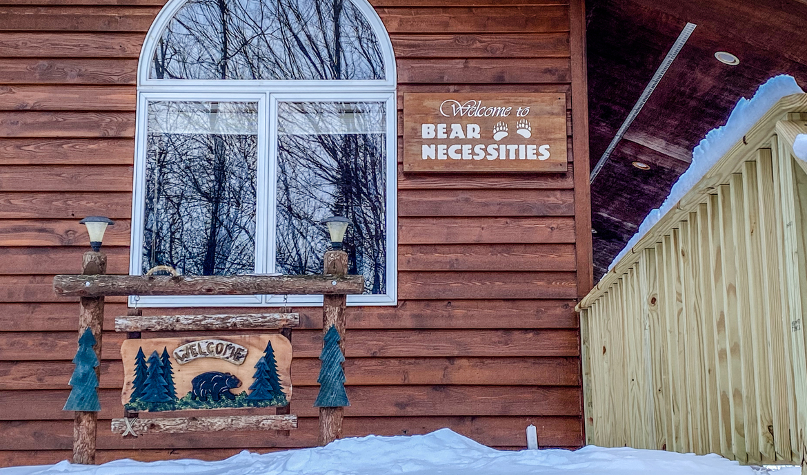 Bear Necessities cabin entrance with snow, wooden welcome sign, and arched window reflecting trees.