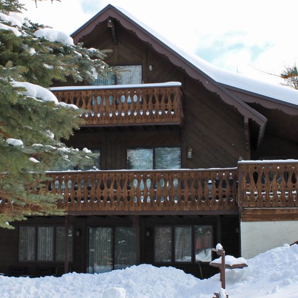Snowy view of Pine Haven cabin with wooden balconies and A-frame roof.