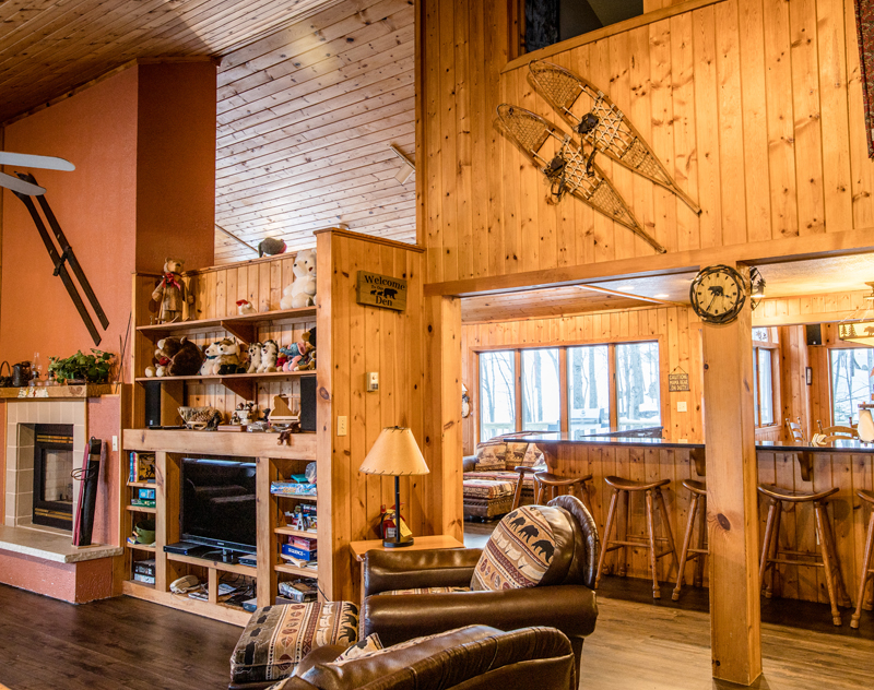 Rustic cabin interior with wood paneling, a stone fireplace, and snowshoes on the wall. "Welcome to the Bear Den" sign visible.