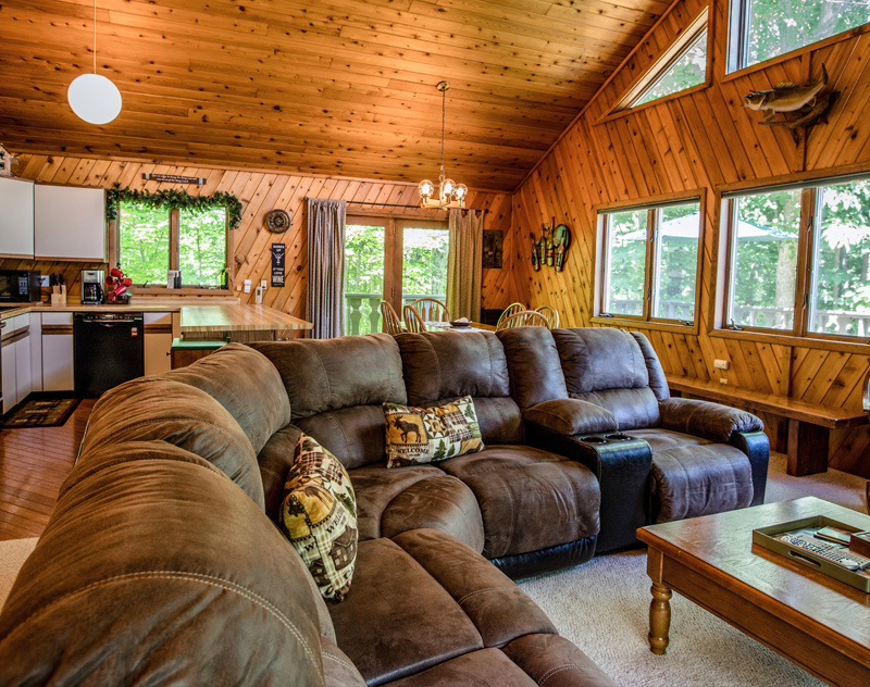 Cozy cabin living room with brown sectional sofa, wood paneled walls and ceiling, and large windows.